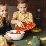 Positive little girl and boy mixing cutted ingredients for salad in big bowl while cooking at table with assorted fresh vegetables and fruits